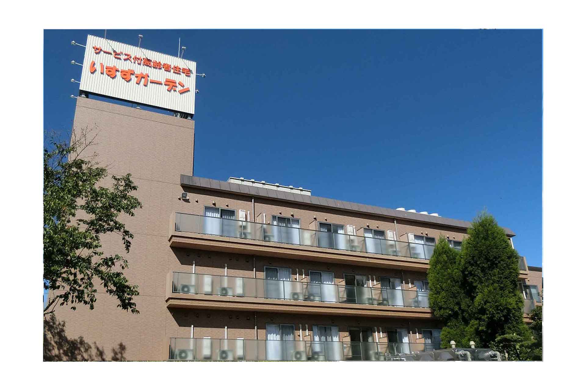 介護施設の建物外観。茶色のタイル張りの3階建て建物で、屋上に日本語の施設名看板が見える。
